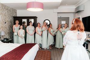 Bride in white lace gown seen from behind facing bridesmaids in sage green dresses holding bouquets in elegant hotel room.