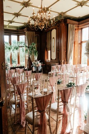 Elegant wedding ceremony room with pink sashed chiavari chairs, chandelier, and floral arch in a grand wood-paneled venue.