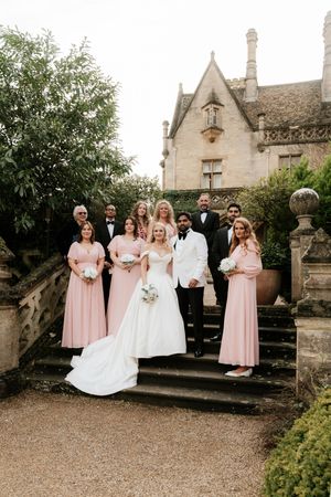 Wedding party poses on stone steps outside a historic manor, bride in white gown with bridesmaids in pink dresses.