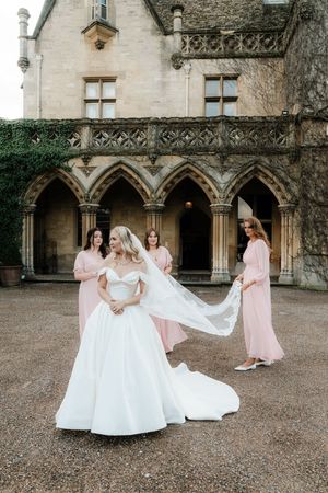 Bride in white off-shoulder gown with veil held by bridesmaids in pink dresses outside a historic stone manor.