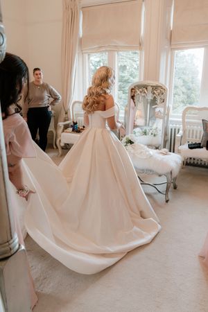 Bride in elegant off-shoulder ballgown wedding dress with long train, viewed from behind in a bright dressing room.