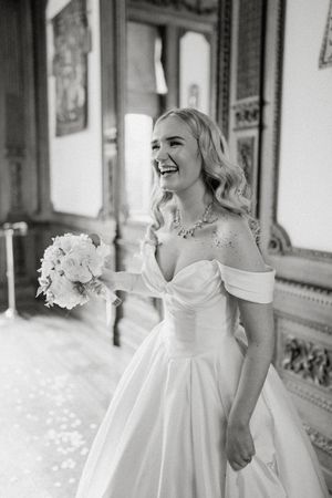 Radiant bride in off-shoulder white gown holding floral bouquet, laughing joyfully in elegant venue, black and white photo.