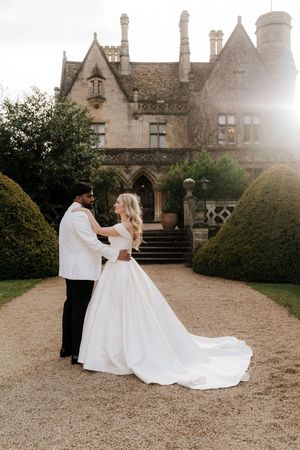 Bride and groom embracing on gravel path before a stunning English manor house at golden hour.