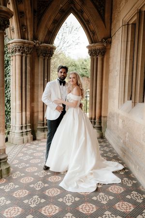 Bride and groom pose elegantly under Gothic stone arches on ornate tiled floor, she in off-shoulder gown, he in white tuxedo.
