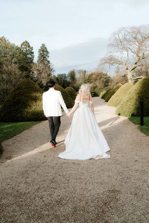 Bride and groom holding hands walking along a garden path, bride in a flowing white ballgown wedding dress.