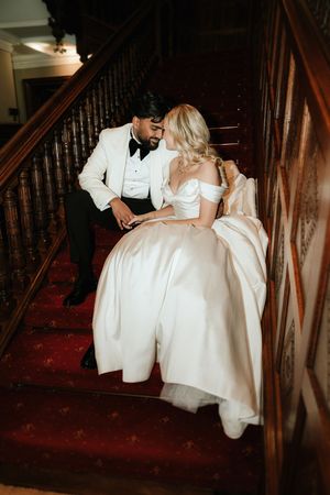 Bride and groom sharing a romantic moment on grand wooden staircase with red carpet in elegant venue.