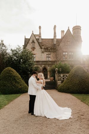 Bride and groom embrace on a gravel path before a grand English manor house with manicured hedges.