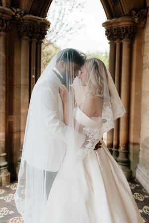 Bride and groom share an intimate moment under a wedding veil at a stone archway venue.