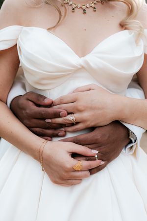 Interracial couple embracing at wedding, showing gold rings, bracelet, and green gemstone necklace on bride in white gown.