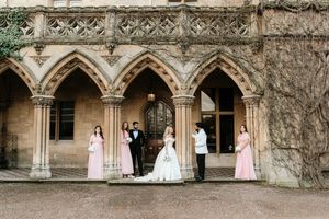 Wedding party poses under Gothic stone arches at historic venue, bride in white gown with pink bridesmaids.