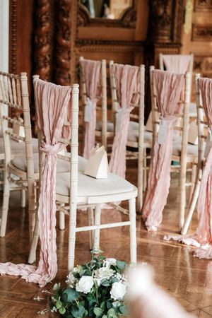 Elegant white Chiavari chairs adorned with flowing pink sashes at a wedding ceremony venue with floral arrangements.