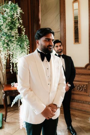 Groom in white tuxedo jacket with black bow tie stands elegantly at wedding ceremony with groomsman in background.