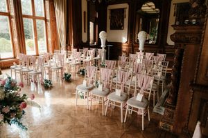 Elegant wedding ceremony setup with white Chiavari chairs adorned with pink ribbons in a grand historic venue.