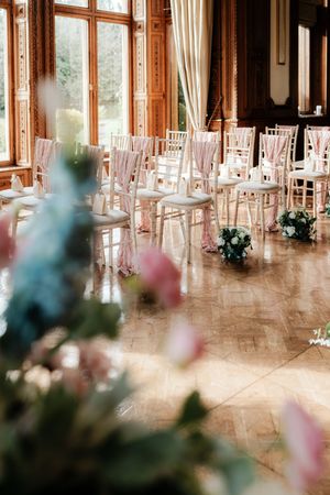 Elegant wedding ceremony venue with white Chiavari chairs, pink ribbon accents, floral arrangements, and sunlit parquet floors.