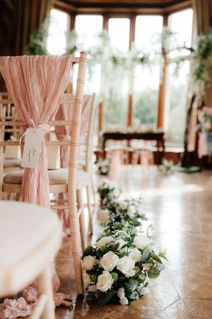 Elegant wedding ceremony aisle with chiavari chairs adorned with pink sashes and white rose floral arrangements on marble floor.