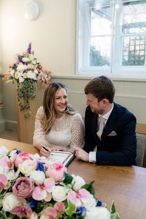 A couple signs documents at a table decorated with pink and white floral arrangements during an indoor ceremony.