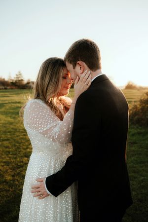 A romantic couple embraces in a sunlit field at golden hour wearing formal attire.