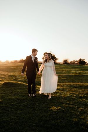 A bride and groom walk together across a sunlit grassy field during golden hour at sunset.