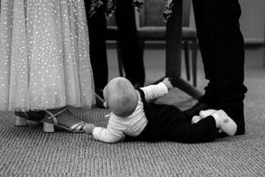 Black and white photo of a baby crawling on carpet near formal attire during a wedding event.