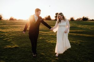 A couple walks hand in hand through a golden sunset field in romantic wedding attire.