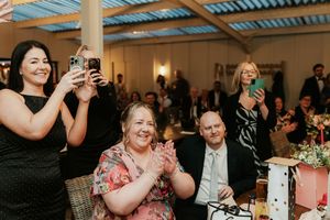 Guests smile and clap at a wedding reception, with some capturing moments on smartphones in an elegant venue.