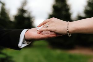 Couple holding hands outdoors, woman wearing diamond engagement ring and silver bracelet, man in dark suit.