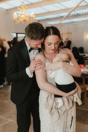 Bride and groom in wedding attire tenderly holding a sleeping baby at their wedding reception.