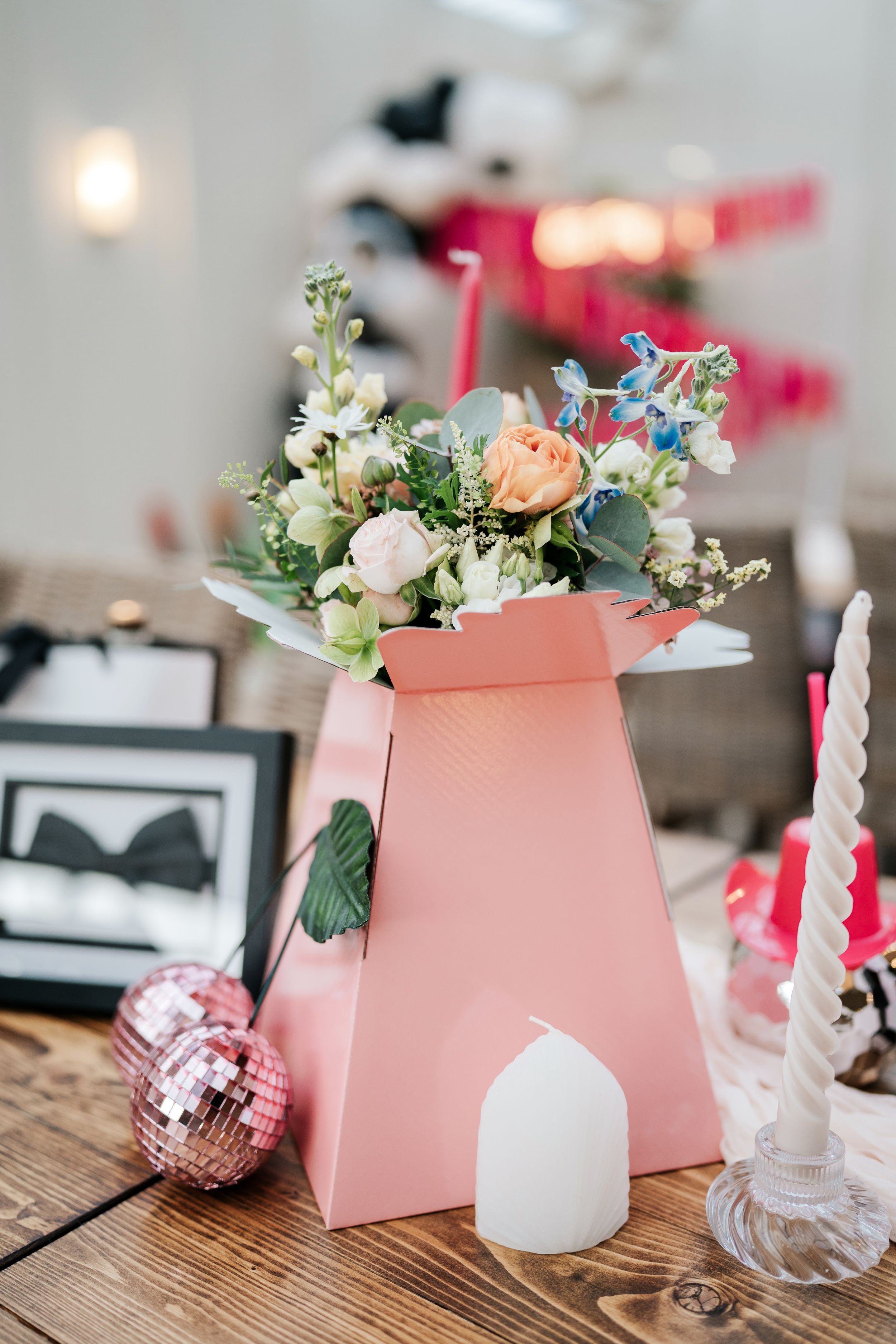 Pink floral arrangement in a decorative box with candles and disco balls on a rustic wooden table.