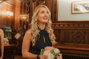 Elegant bridesmaid in navy dress holding pink bouquet in wood-paneled room.
