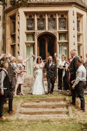 Wedding party and guests throw confetti outside a Gothic-style mansion.