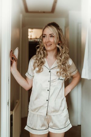Woman in white silk pajamas poses playfully in a bright hallway during wedding morning preparations.