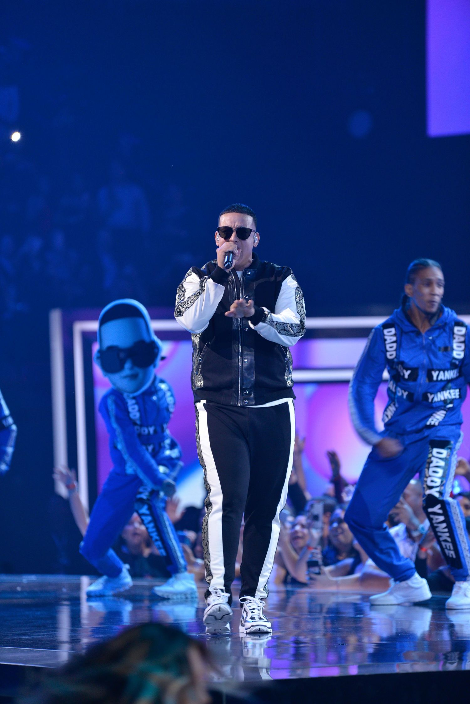 Performer in black vest and sunglasses singing on stage with dancers in blue costumes.