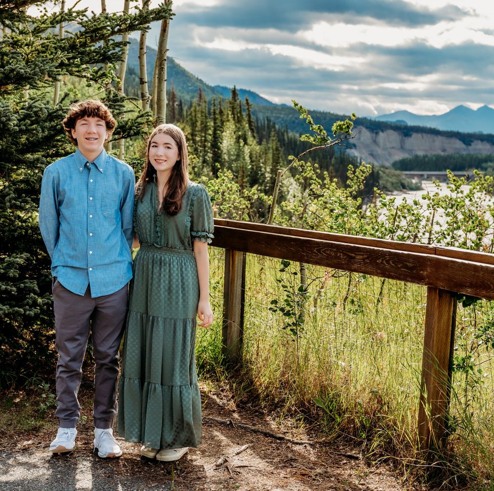 Couple standing together on mountain overlook with dramatic cloudy sky and evergreen forest.