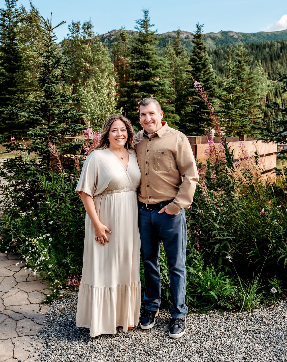 Couple in casual attire posing in mountain garden setting with evergreen trees.