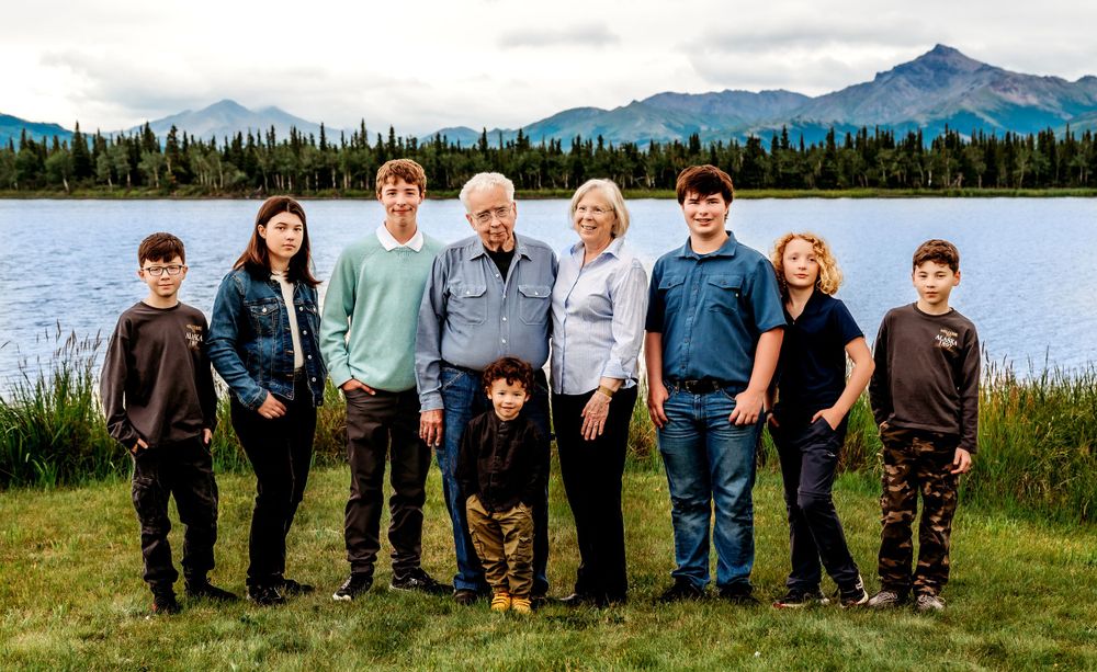 Family with dog gathered by lakeside with snow-capped mountains in background.