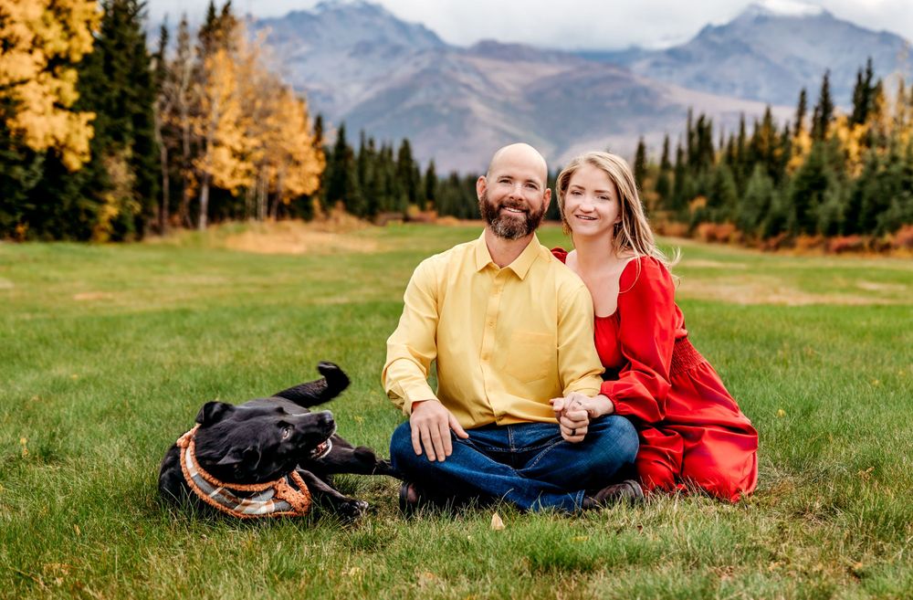 Couple sitting on grass with black dog against mountain landscape and autumn trees.
