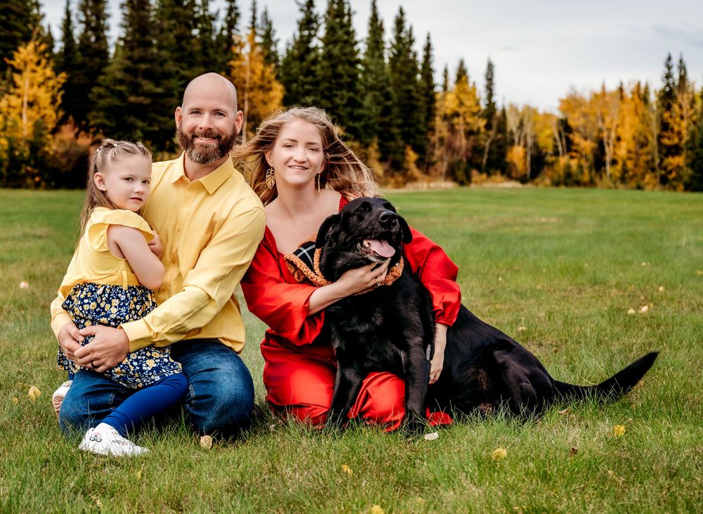Family with black dog poses together on grass with autumn mountain landscape.