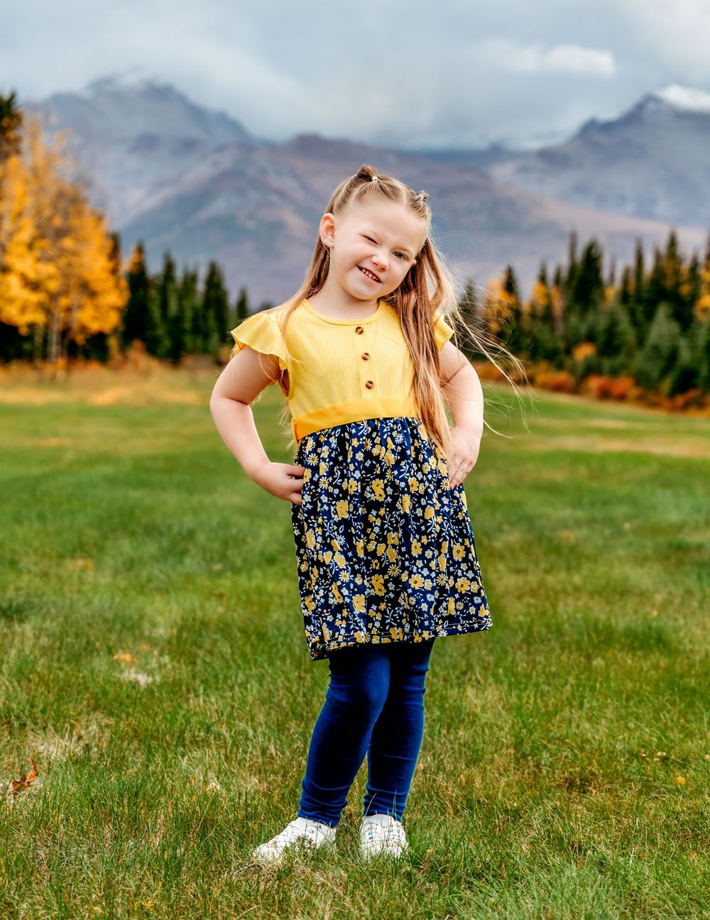 Young girl in yellow top and floral skirt poses joyfully against mountain landscape.