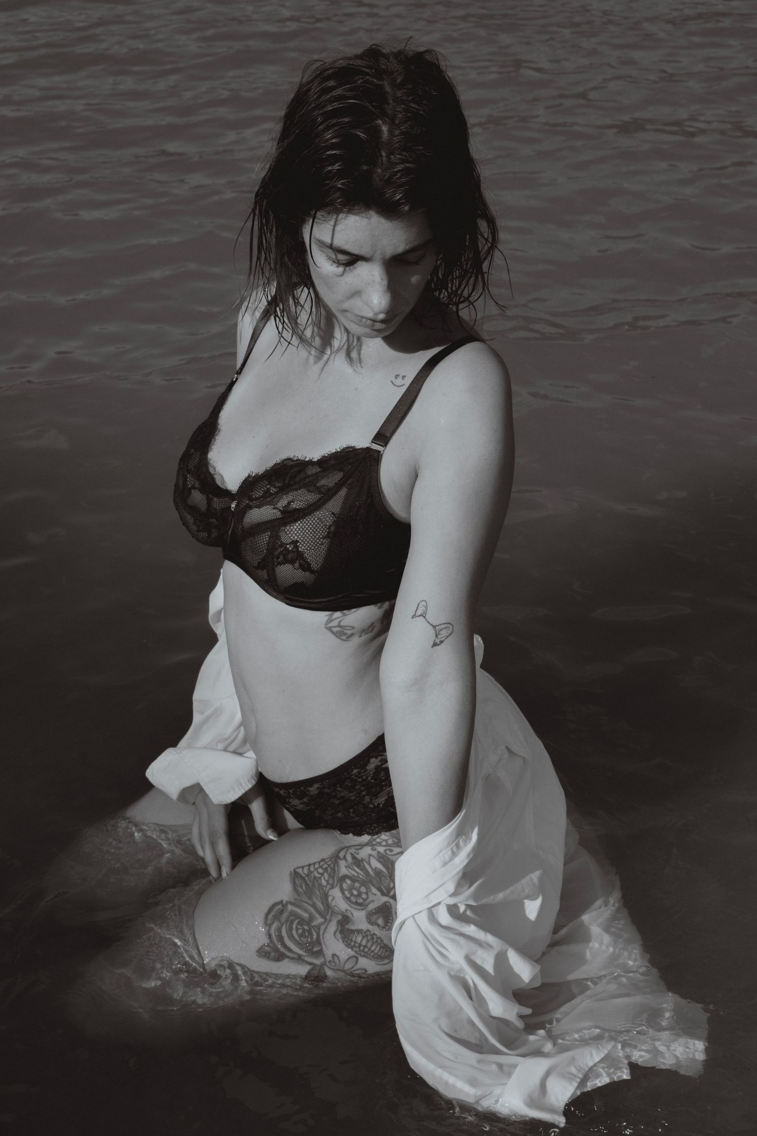 Woman in black bra and white shirt standing in a lake with a rose tattoo on her thigh, mountains in background.