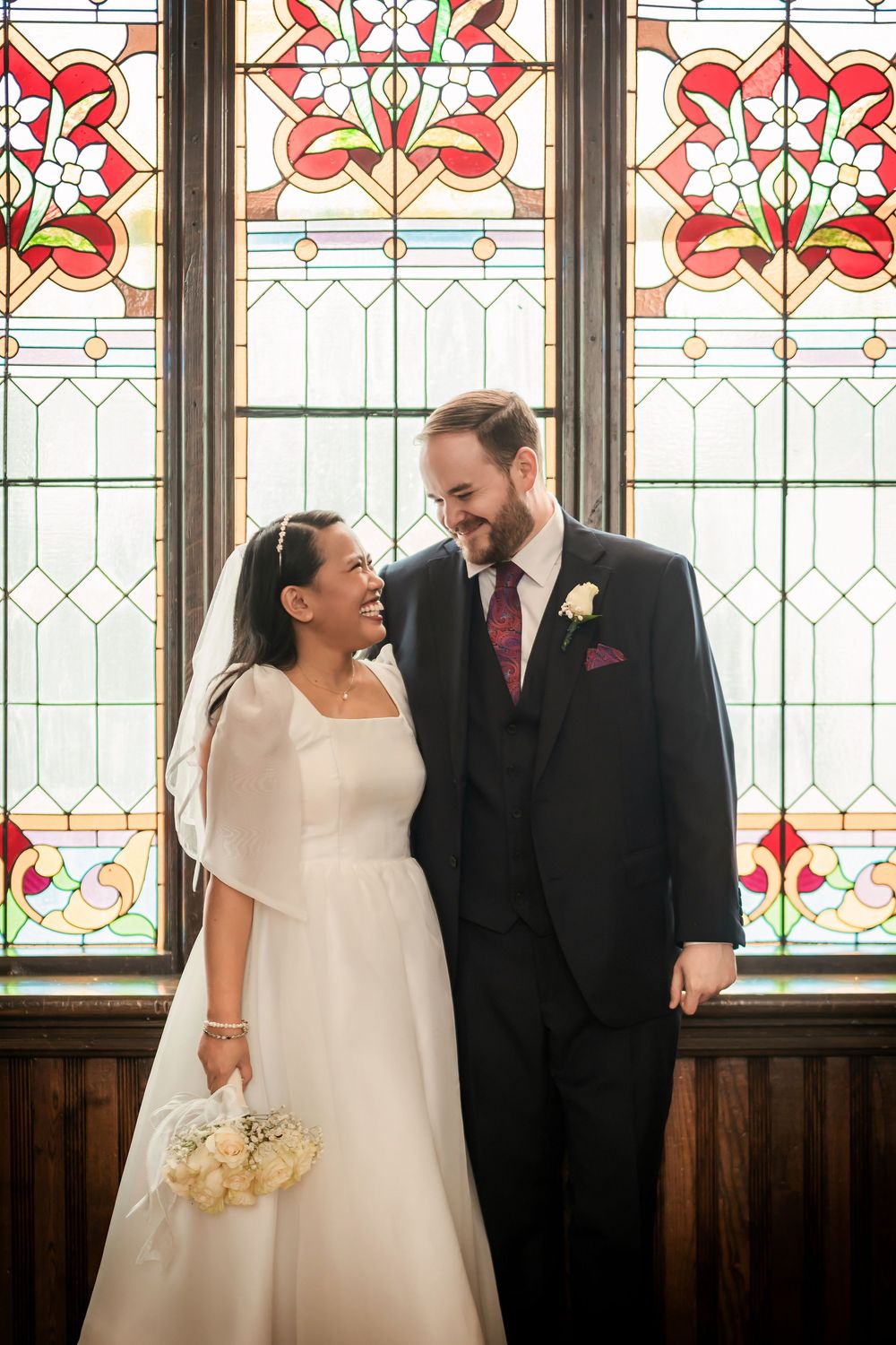 Bride and groom smiling at each other in front of a colorful stained glass church window on their wedding day.
