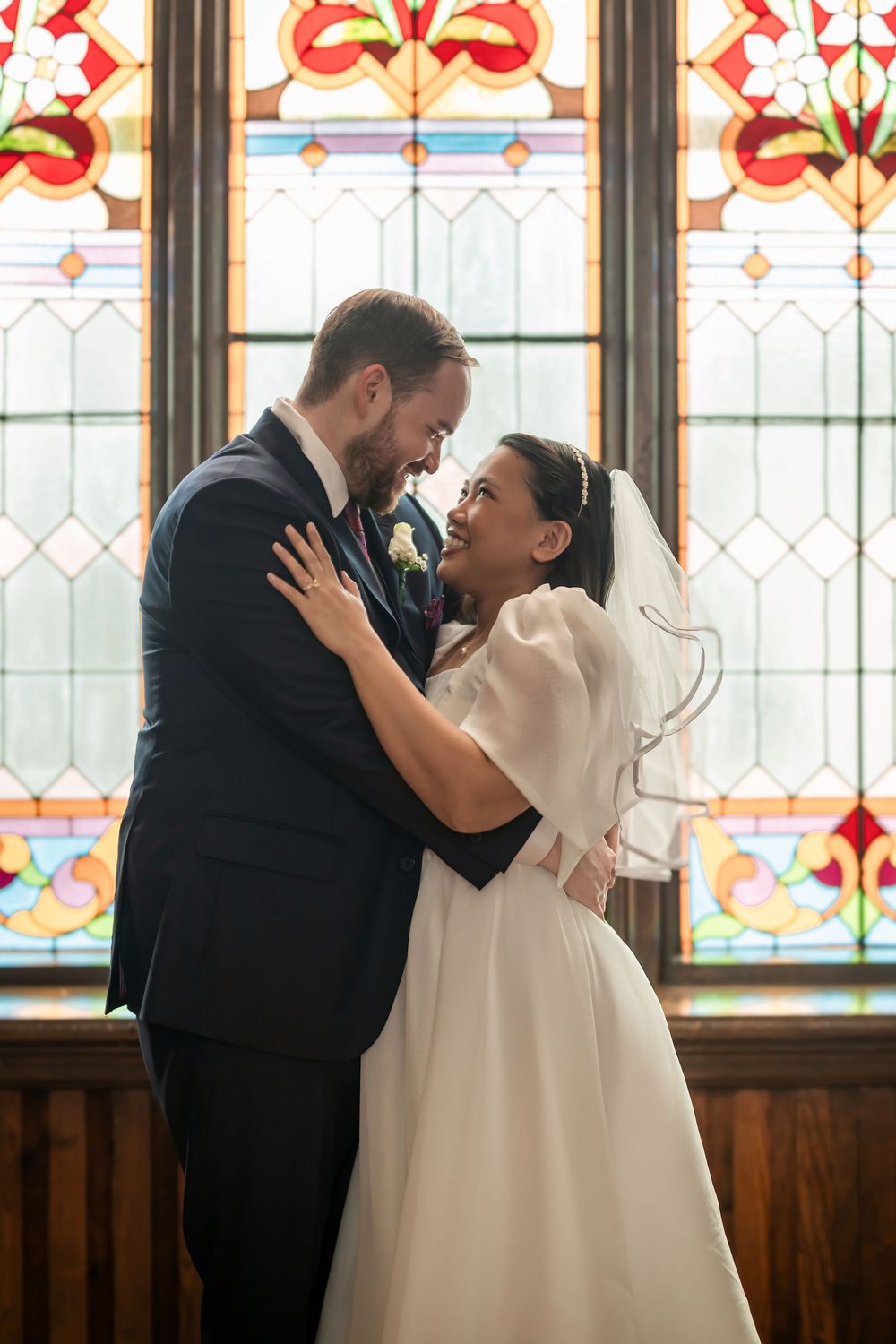 Bride and groom embracing and smiling at each other in front of a colorful stained glass window inside a church.