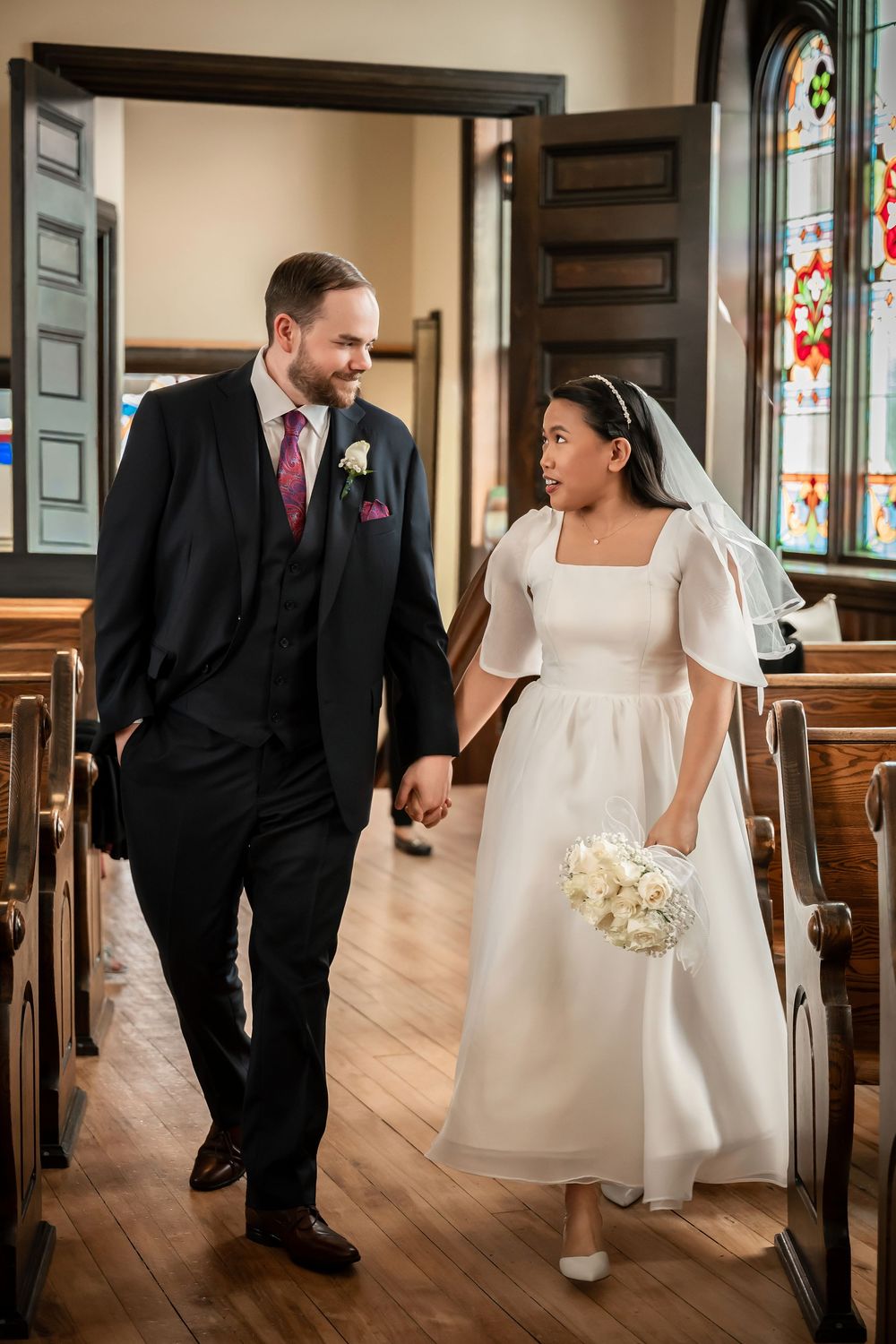 Newlywed couple walking down church aisle, bride in white midi dress holding bouquet, groom in black suit, stained glass windows.