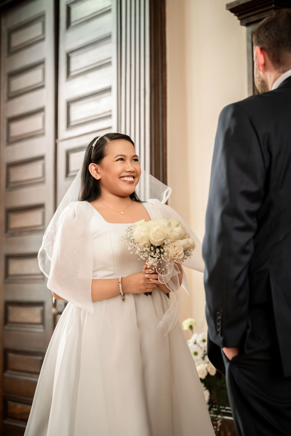 Smiling bride in white gown holding bouquet faces groom in black suit at church doors on wedding day.
