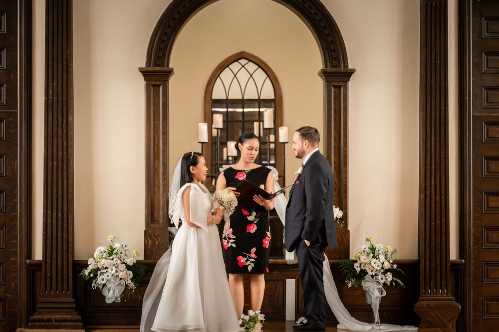 Bride and groom exchange vows with officiant in elegant chapel with arched doorways and floral arrangements.