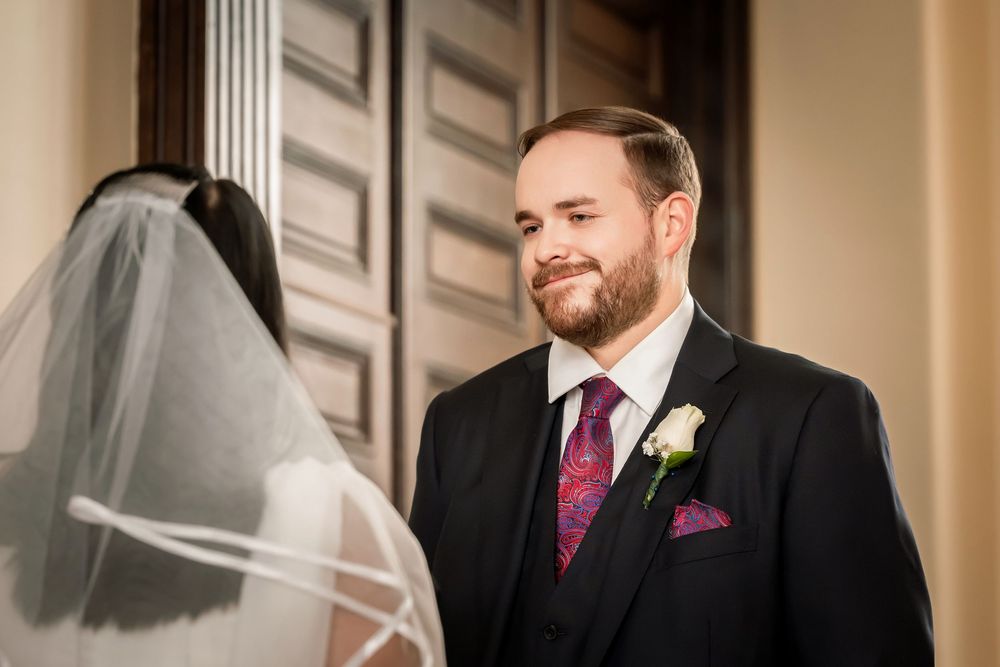 Groom in black suit with purple tie smiles at bride in veil during wedding ceremony first look moment.