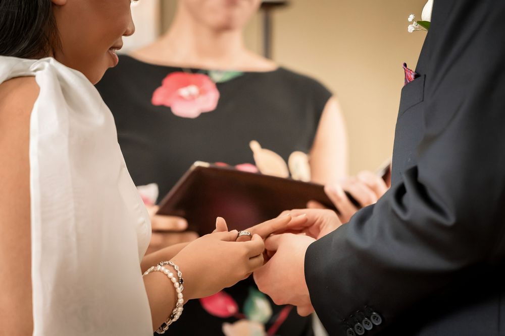Bride placing a wedding ring on groom's finger during ceremony, with officiant holding book in background.