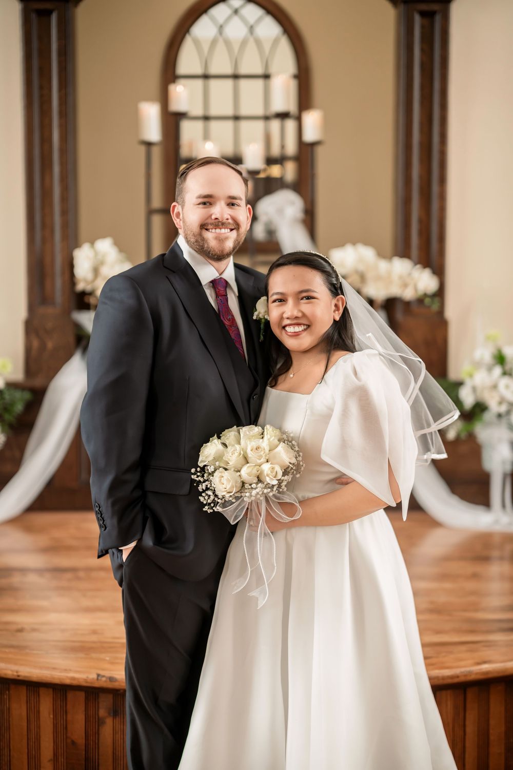Smiling newlywed couple posing in a decorated church, bride in white gown holding bouquet, groom in dark suit.