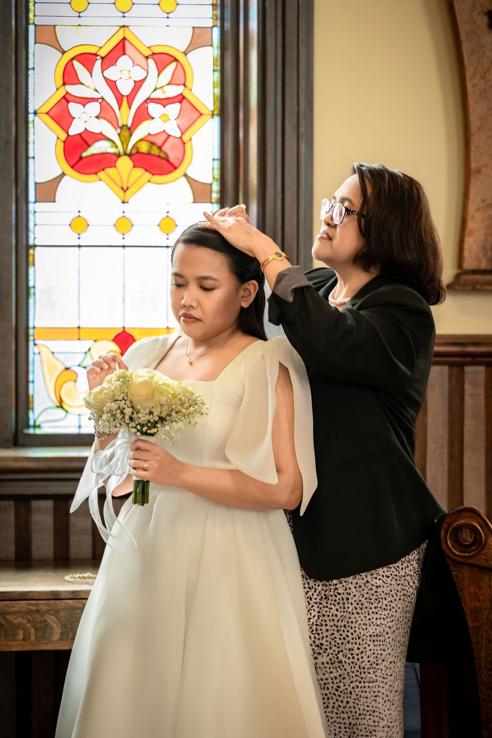A bride in a white gown holds a bouquet while a woman adjusts her veil inside a church with stained glass windows.
