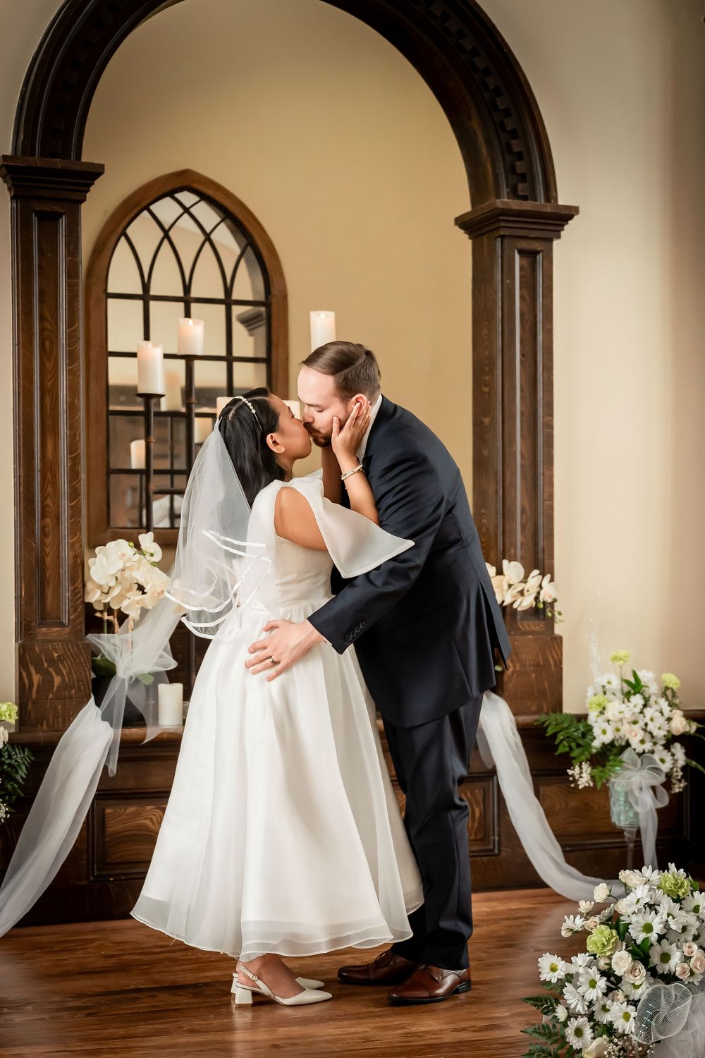 Bride and groom share first kiss at altar in chapel, bride wearing tea-length gown with veil, groom in navy suit.