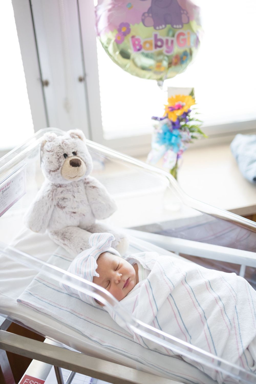 Newborn baby girl sleeping in hospital bassinet with teddy bear and Baby Girl balloon during her Fresh 48 session at Fort Sanders.